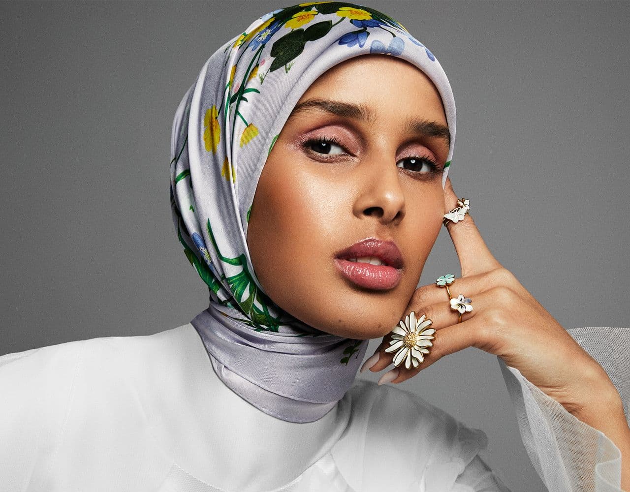 Portrett av Rawdah Mohamed, som ser rett inn i kameraet. Hun har på seg flere ringer med emaljeblomster i ulike pastelltoner.